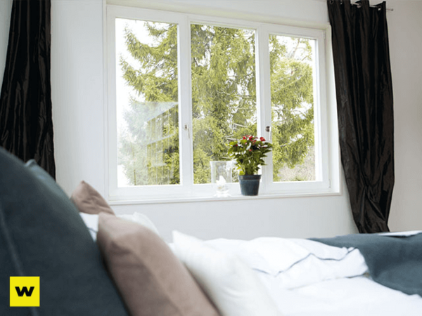 Helles Schlafzimmer mit großem Fenster, gemütlichem Bett und Topfpflanze auf Fensterbank.