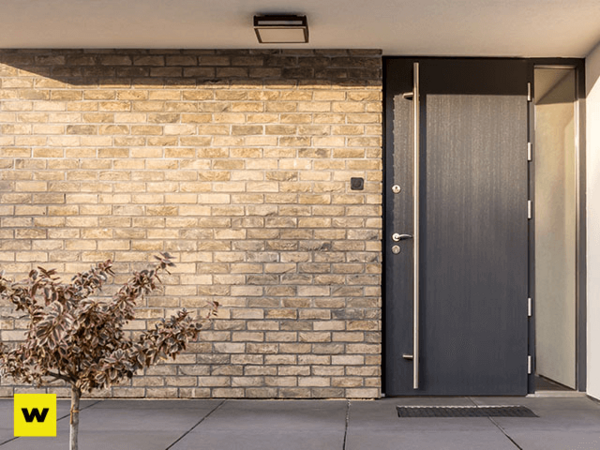 Moderne Haustür aus dunklem Holz mit Glaseinsatz vor heller Klinkerfassade und Pflanze.